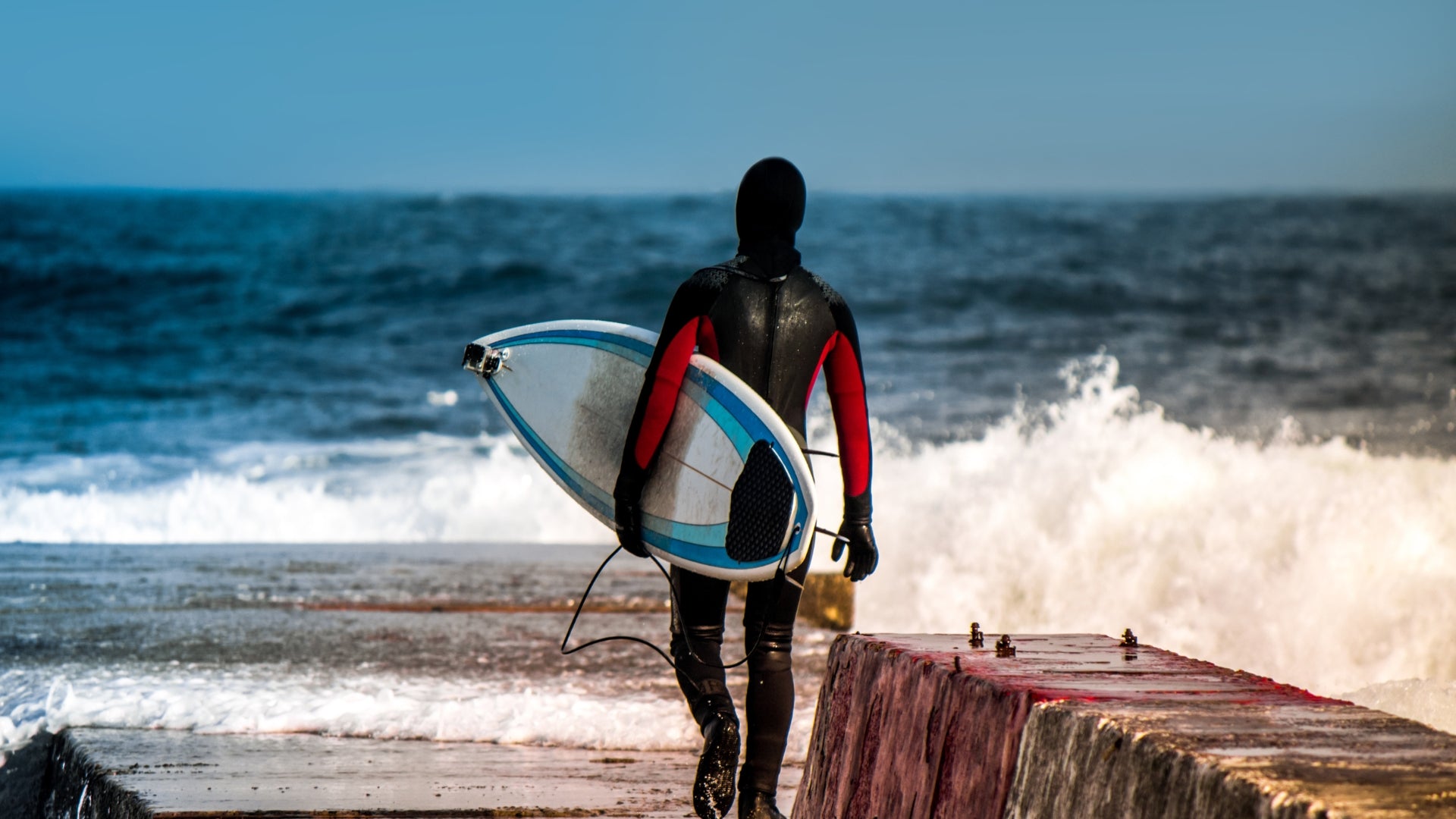 Surfer in a hooded wetsuit carrying a surfboard beside cold ocean waves