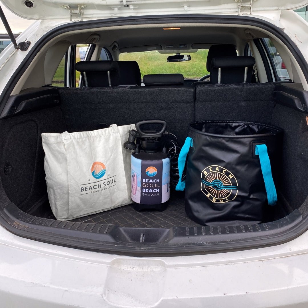 Beach Soul branded beach shower tote bag, portable pressure shower, and collapsible bucket with bright turquoise handles neatly packed in the trunk of a white car