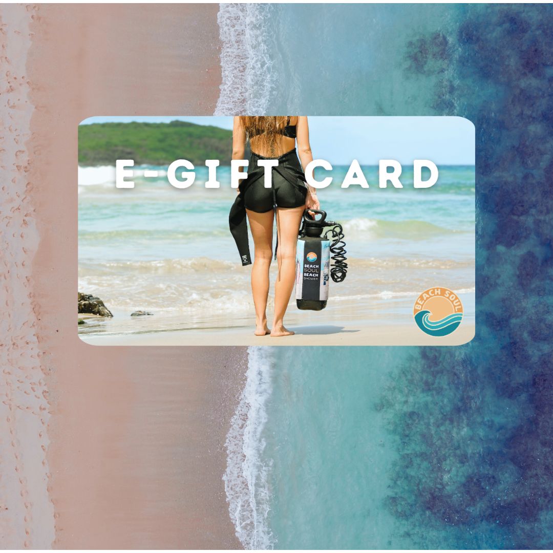Photograph of an e-gift card promotion featuring a woman in a black wetsuit holding a portable Beach Soul shower on a tropical beach with pink sand and clear turquoise water overlaid with E-GIFT CARD text and the Beach Soul logo