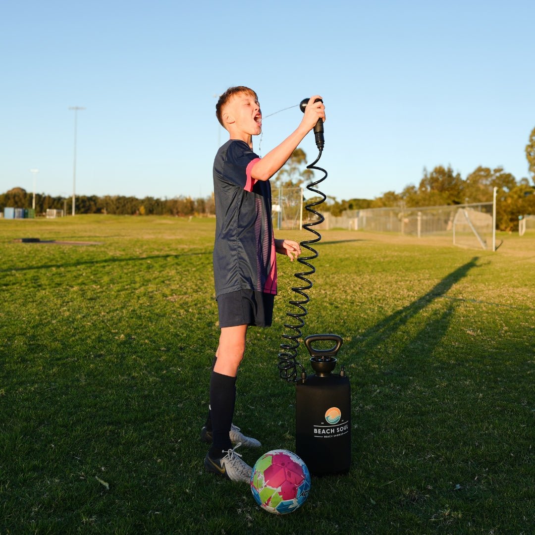 A young boy in a navy and pink soccer uniform takes a refreshing drink from a black Beach Soul portable shower on a bright green sports field, with a vibrant multi-colored soccer ball at his feet and a distant soccer goal visible