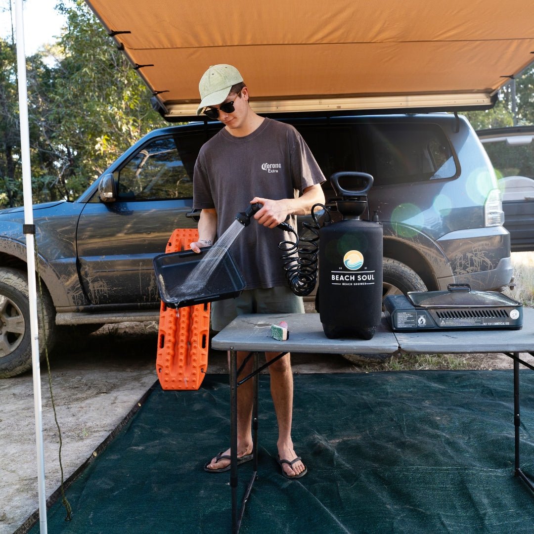 A man in a Corona Extra t-shirt and green cap rinses a black pan using a Beach Soul portable shower system at an outdoor campsite beside a dirty grey SUV with orange recovery tracks and a camping stove on a grey table