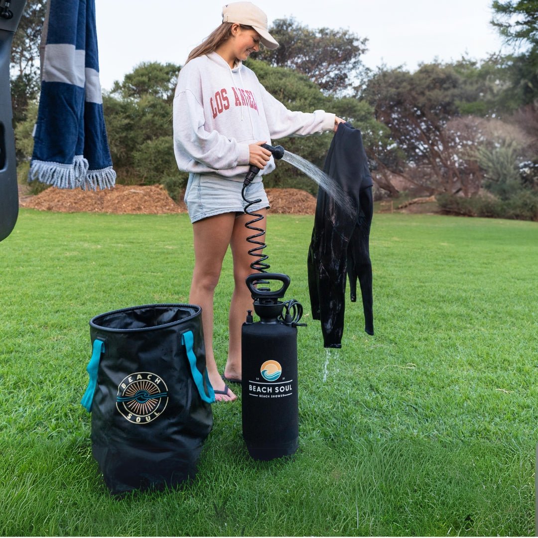A young woman in a beige cap and Los Angeles sweatshirt rinses a black wetsuit with a Beach Soul portable shower next to a matching bucket with teal handles on a grassy outdoor area