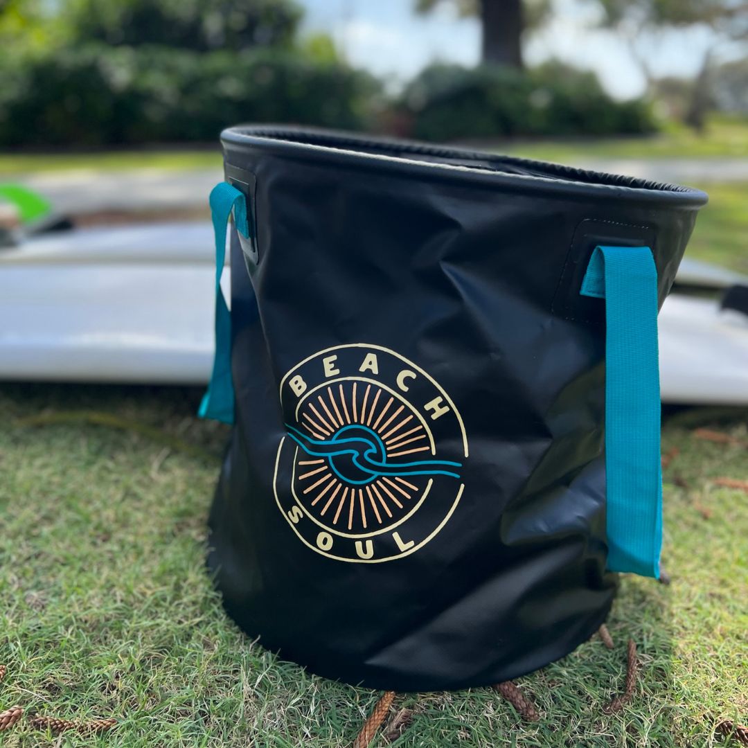 Black flexible bucket bag with teal handles showcasing a BEACH SOUL logo featuring a sun and wave design, placed on green grass with blurred white surfboards behind it