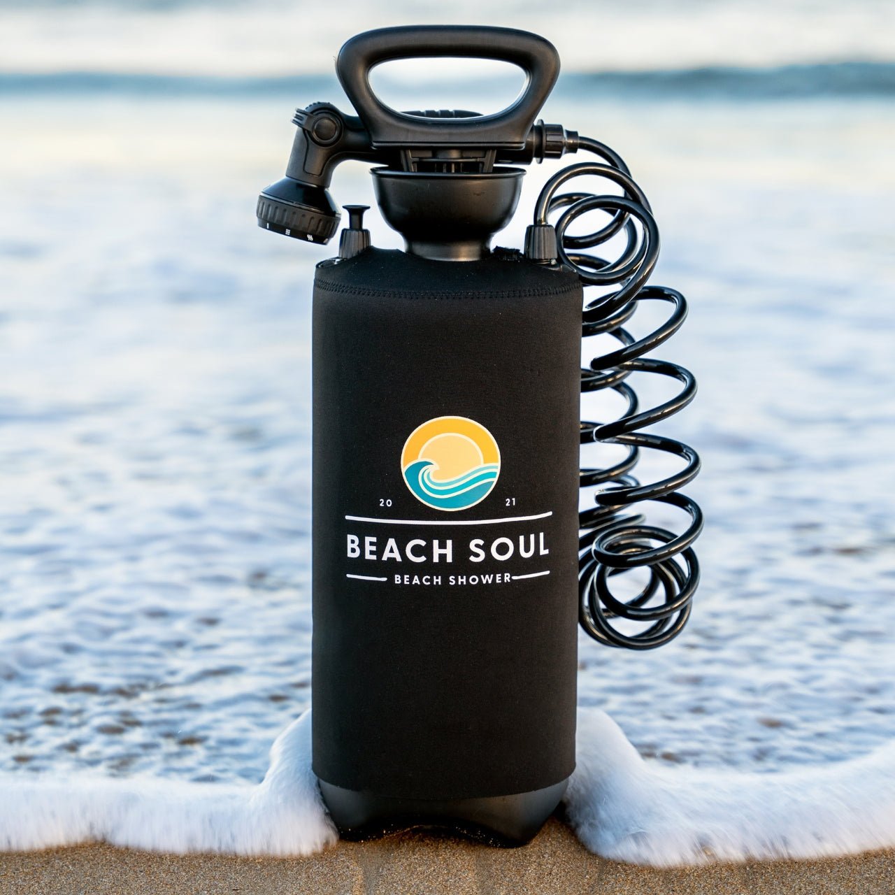 Black Beach Soul portable shower with a vibrant sun and wave logo, coiled hose, and showerhead standing in frothy ocean water on a sandy beach
