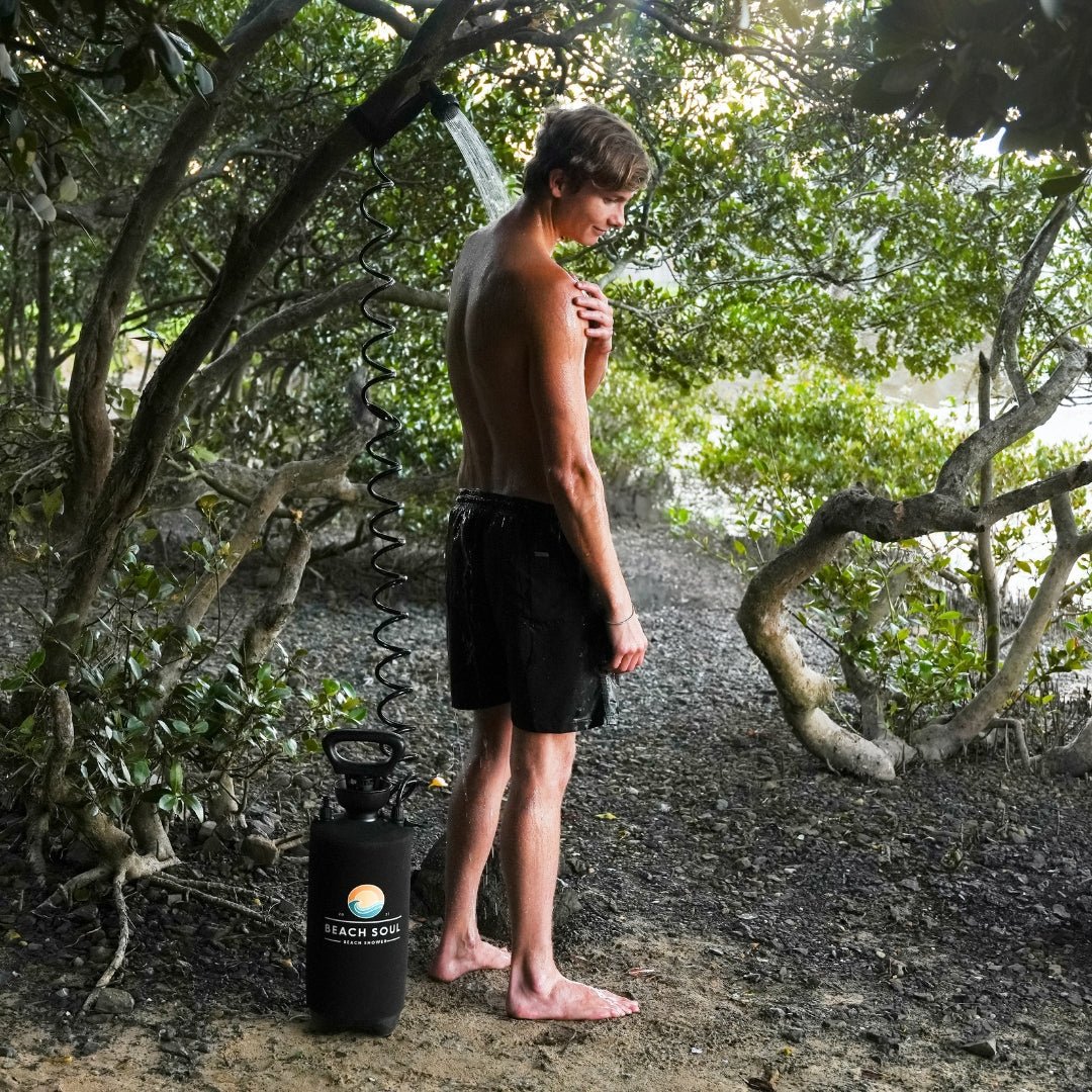 Photograph of a young man with wet hair and black shorts showering outdoors using a portable black Beach Soul shower unit with a colorful logo, water spraying, surrounded by lush green foliage and visible tree roots on a sandy path