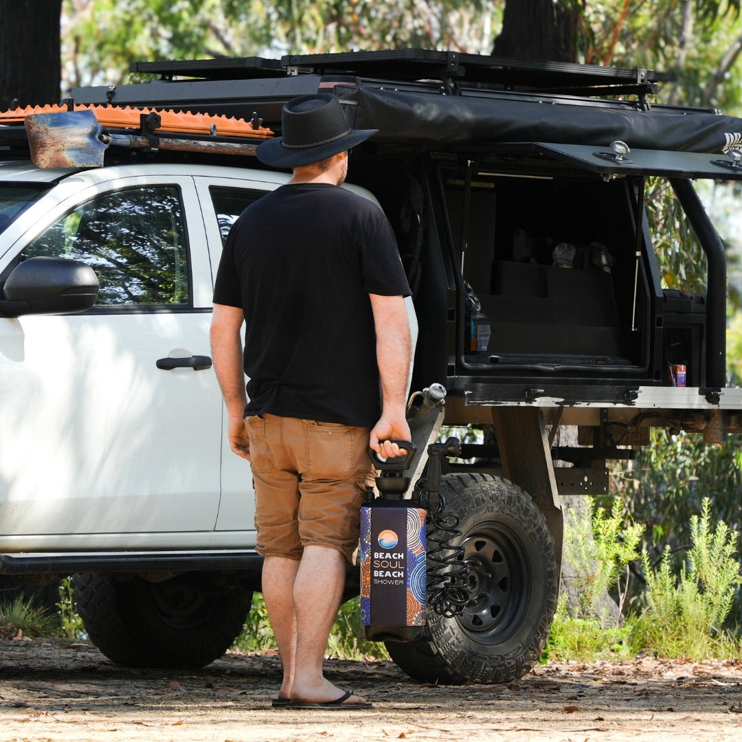 A man in a black hat stands beside his white off-road pickup truck with a black roof rack and shovel, holding a portable Beach Soul Beach Shower with a colorful wave and dot pattern in a natural wooded setting