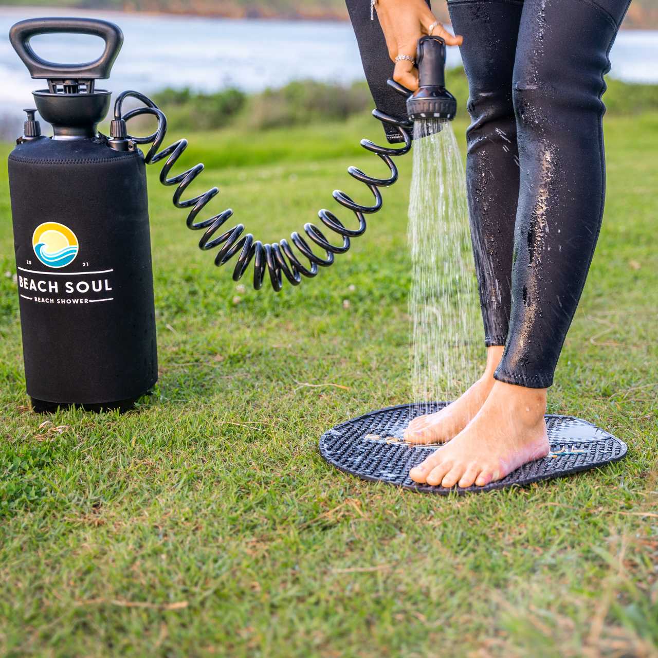 Photograph of a person in black wetsuit pants rinsing their bare feet on a black textured mat with a Beach Soul portable shower tank featuring a yellow sun and blue wave logo on a grassy beach shore