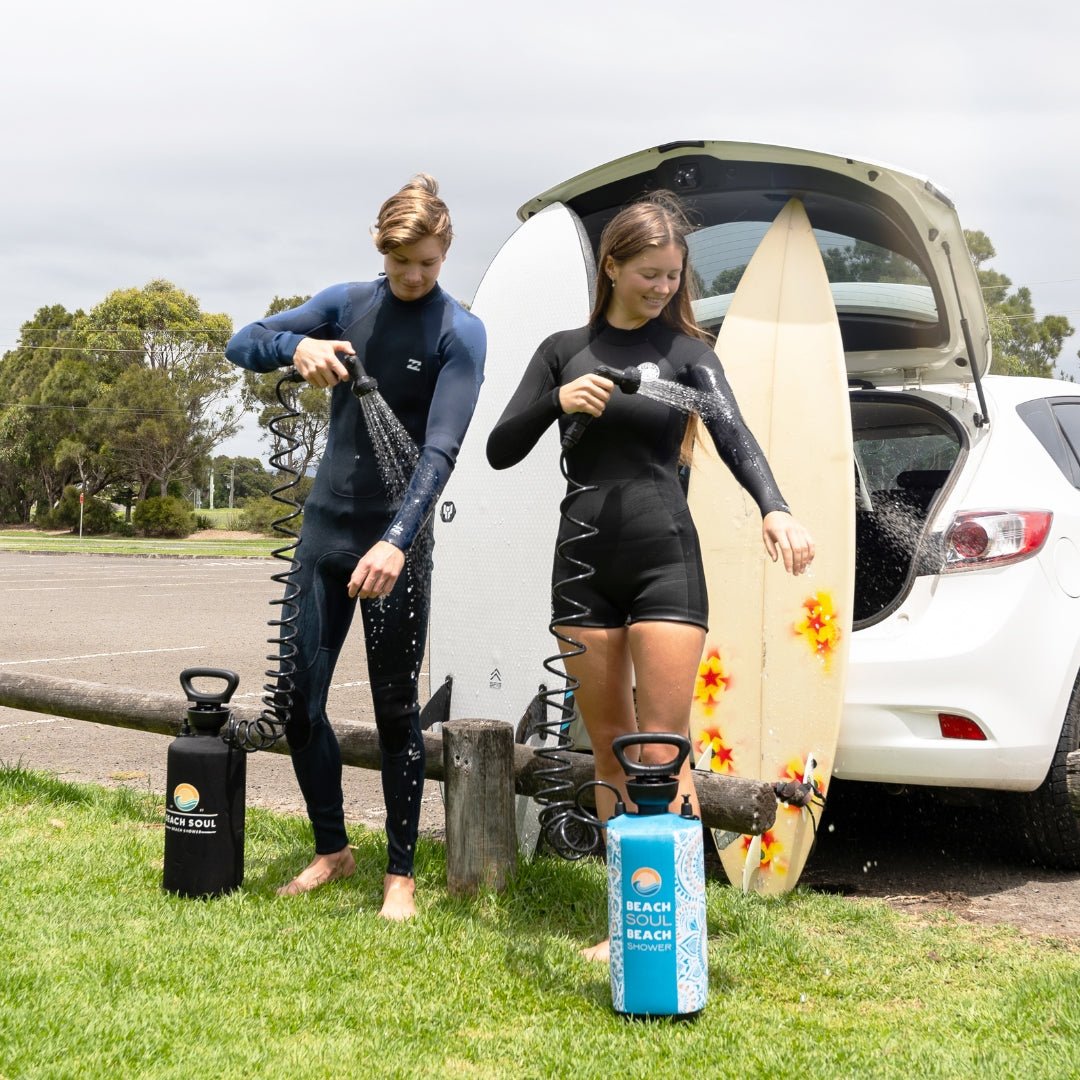 Two surfers using Beach Soul portable showers to rinse their wetsuits after surfing