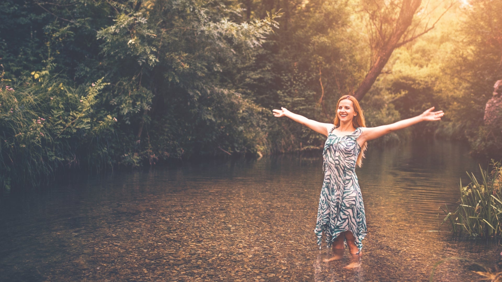 Woman standing in a river with arms outstretched, surrounded by greenery