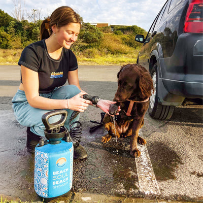 A smiling young woman in a Boards & Bands crop top and light blue sweatpants gently washes a brown dog's paw using a blue portable Beach Soul Beach Shower next to a black SUV on a paved area