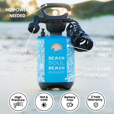 Product photography of a Beach Soul Beach Shower, a portable unit featuring a bright blue insulated cover with a white and orange mandala pattern, a distinctive sun and wave logo, and a black coiled hose with a 10-mode showerhead, set on a sandy beach at sunset
