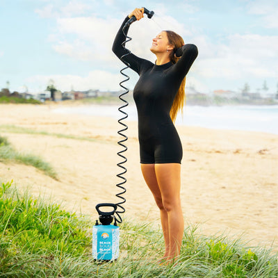 A woman in a black shorty wetsuit joyfully rinses off with a portable blue Beach Soul shower and coiled hose on a golden sandy beach with green grass, ocean waves, and distant coastal housing under a blue sky