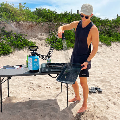 A young man on a sandy beach washing a dark cooking tray with a blue Beach Soul portable shower next to a camping stove and green dune vegetation, wearing a Happy Days cap and sunglasses