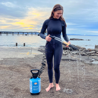 A young woman in a black wetsuit stands barefoot on a gravel beach using a blue portable Beach Soul Beach Shower with water spraying, a long pier visible across the ocean under a colorful sunset sky