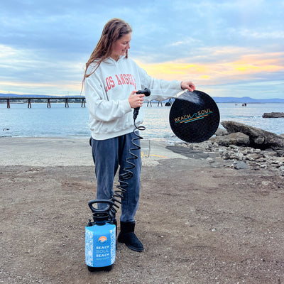 Photography of a smiling young woman in a white LOS ANGELES hoodie holding a black BEACH SOUL round mat and shower nozzle, next to a blue BEACH SOUL portable shower on a rocky shore with a distant pier under a soft sunset sky