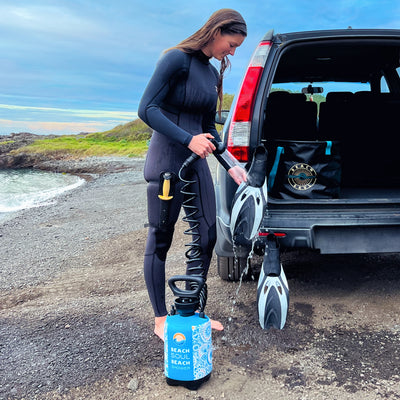 A woman in a black wetsuit rinses black and white diving fins using a blue Beach Soul portable shower beside a grey SUV at a rocky beach with green hills and a branded Beach Soul bag visible in the open trunk