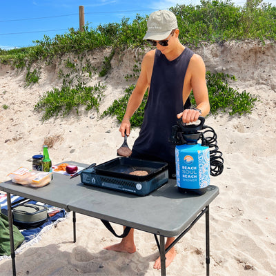 A photograph shows a man wearing a "Happy Days" cap cooking burgers on a portable grill at a sandy beach with a blue "Beach Soul Beach Shower" and fresh ingredients on a folding table backed by green dune vegetation