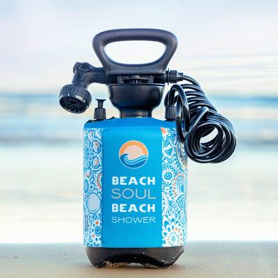 A portable Beach Soul Beach Shower with a blue and white patterned sleeve featuring an orange sun and ocean wave logo, black handle, adjustable nozzle, and coiled hose sits on wet sand at a beach with the ocean in the background