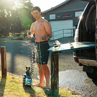 Photography of a shirtless man in a black cap rinsing a blue surfboard with a portable camouflage shower, near a black SUV at The Shellharbour Club and Warilla Bowls, featuring sunlight catching water spray and distinct striped surfboard fins
