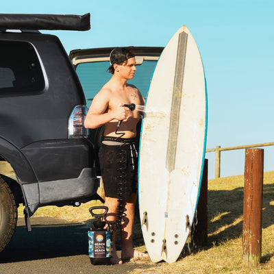A shirtless man in a black mesh cap washes a sandy surfboard with a colorful portable Beach Soul shower next to a dark grey SUV on a sunny grassy beach overlook