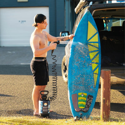 A shirtless man in a black cap washes a sandy blue surfboard featuring yellow geometric patterns with a Beach Soul Shower hose next to an open SUV in a sunlit coastal parking lot