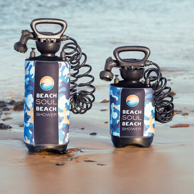 Two portable Beach Soul Beach Shower devices with blue and tan camouflage sleeves and black coiled hoses stand on a sandy beach next to shallow water, each featuring an orange sun and blue wave logo