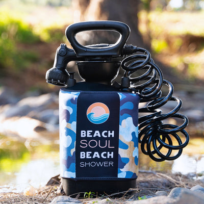 Photograph of a black portable Beach Soul Beach Shower with a blue and white camouflage pattern sleeve featuring an orange sun and blue waves logo, a black coiled hose and shower head, standing on rocks next to a stream outdoors