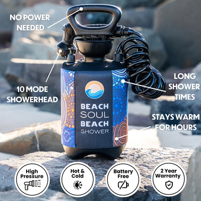 A portable Beach Soul Beach Shower product displayed on a rocky outdoor surface, featuring a black handle, coiled hose, and a vibrant blue and orange patterned sleeve with aboriginal-style designs, highlighting its 10-mode showerhead and no power needed operation