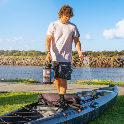 Photograph of a man using a colorful Beach Soul Beach Shower to spray water onto a dark green kayak by a river with a rocky breakwater under a bright blue sky