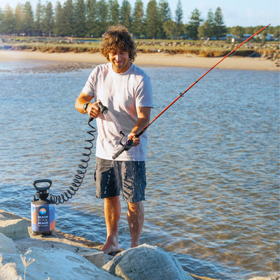 Digital photograph of a smiling man with curly hair rinsing an orange fishing rod with a colorful Beach Soul portable shower on a sandy riverbank with pine trees in the background