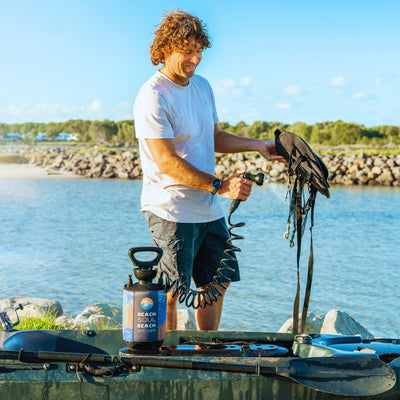 A smiling man with curly hair uses a "Beach Soul Beach Shower" portable sprayer to clean black equipment straps over a green kayak, standing by a blue river with a rocky bank and distant trees under a clear blue sky