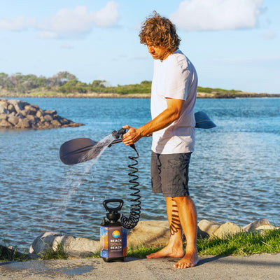 Photography of a man with curly brown hair cleaning a dark paddle with a portable Beach Soul Beach Shower featuring a colorful patterned sleeve and coiled black hose, standing barefoot beside a calm blue estuary with a rocky breakwater under a bright sky