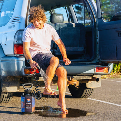 A smiling man with curly hair sits on the open tailgate of a silver 9400V6 Quad Cam SUV, using a colorful Beach Soul portable shower to rinse his bare feet in a sunlit parking lot