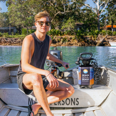 A young man in sunglasses and a black tank top sits in an aluminum boat featuring an outboard motor, alongside a portable 'Beach Soul Beach Shower' with vibrant Indigenous-inspired art, on calm blue water bordered by lush green trees