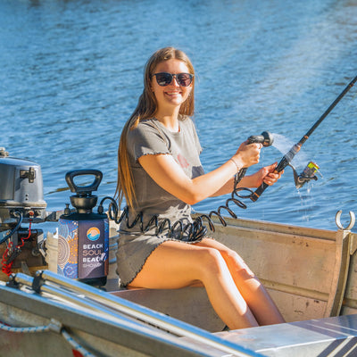 A young woman in sunglasses smiles in a metal boat on calm blue water, using a portable Beach Soul Beach Shower with its unique blue, orange, and white graphic design to rinse a fishing rod with bright yellow line