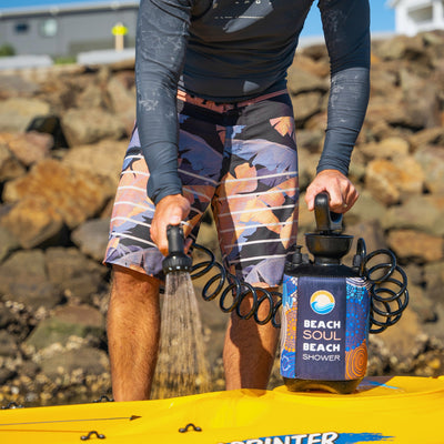 A man in a dark rash guard and vibrant tropical board shorts sprays water from a black Beach Soul Beach Shower with a colorful patterned sleeve, resting on a bright yellow kayak at a rocky shoreline