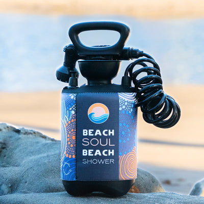 A black portable Beach Soul beach shower with a colorful blue, orange, and white patterned neoprene sleeve featuring a sun and wave logo and a coiled hose rests on a sandy beach with the ocean in the background