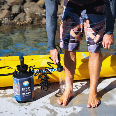 A person wearing a grey rash guard and colorful tropical print board shorts showers their bare feet with a portable Beach Soul Beach Shower, which has a blue and orange wave logo and aboriginal-style patterns on its neoprene sleeve, standing on a sunny concrete dock beside a bright yellow kayak by calm water