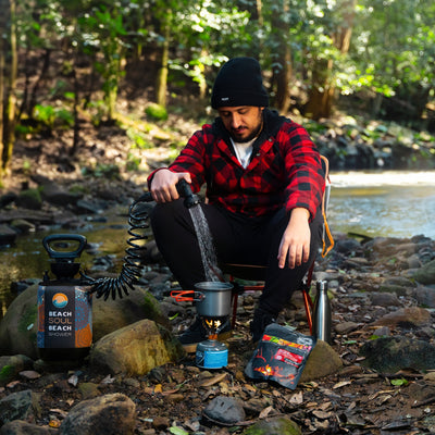 A man in a red plaid shirt and black beanie pours water from a portable Beach Soul Beach Shower into a camping pot on a stove by a river in a green forest, with a packaged The Green Chicken Curry meal nearby