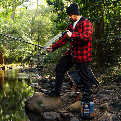 Photograph of a man in a red and black plaid jacket and black beanie cleaning a fishing rod with a colorful portable Beach Soul Shower at a rocky forest stream surrounded by lush green foliage