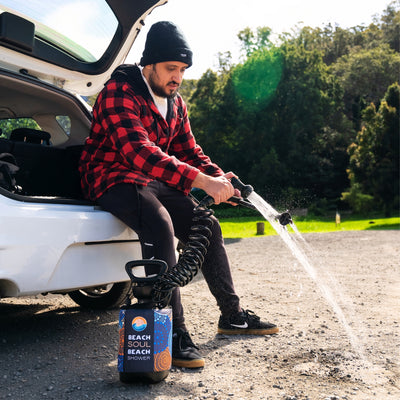 A man in a red plaid shirt and black beanie sits on the open trunk of a white car, spraying water from a portable Beach Soul Beach Shower with its colorful blue and orange design onto the gravel ground, framed by lush green trees
