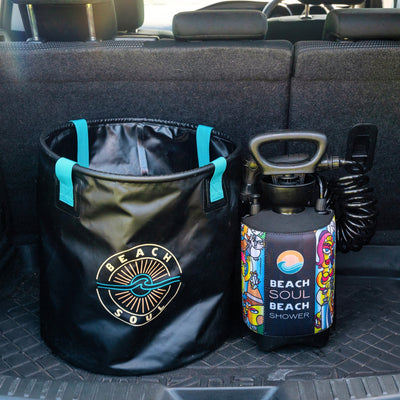 A black Beach Soul collapsible bucket with teal straps and a portable pressure shower with a colorful abstract sleeve by Steven Bormann and a black coiled hose are displayed in a car trunk