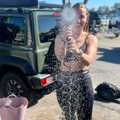 A woman in a black bikini top and dark leggings showers outdoors, holding a coiled hose with water splashing over her smiling face, next to a military green Suzuki Jimny SUV and a light purple tub on the ground