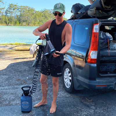 A young person in a green hat and sunglasses uses a blue Beach Soul portable shower to rinse their bare legs, water splashing next to a dark grey Honda camping van laden with gear on its roof, set against a vibrant turquoise lake and lush green trees