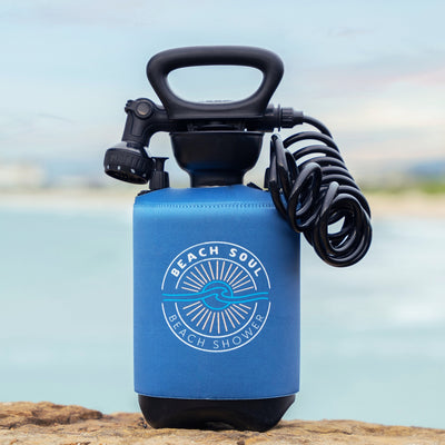 A "Beach Soul Beach Shower" portable pump shower featuring a blue neoprene sleeve with a sun and wave logo, a black pump handle, and a black coiled hose, resting on a rocky shore with a blurred blue ocean in the background