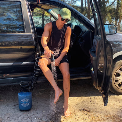 Photography a young man in a green corduroy cap and sunglasses sits in a black SUV's open door washing his sandy bare feet with a portable blue "Beach Soul Beach Shower" spray bottle outdoors near trees and distant water