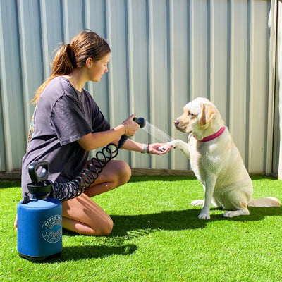 A young woman in a ponytail kneels on bright green artificial turf washing a yellow Labrador retriever's paw with a portable blue Beach Shower sprayer, the dog wearing a vibrant pink collar next to a gray corrugated fence