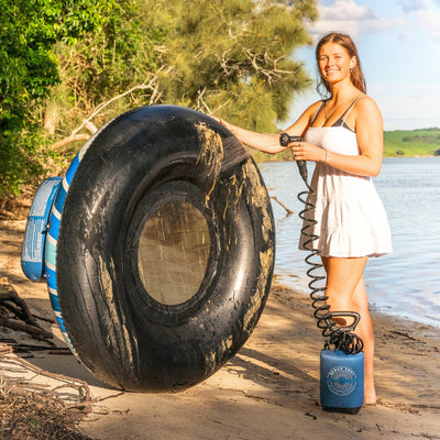 A smiling young woman in a white sundress cleans a sandy black river tube with a visible speaker and blue stripes, using a blue portable "Beach Soul" shower with a coiled hose on a bright riverbank