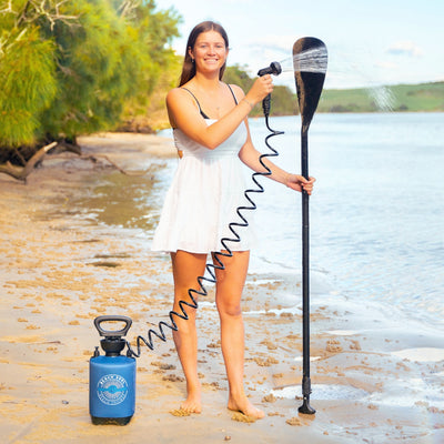 A smiling young woman in a white dress sprays a black paddle with water from a blue Beach Soul Beach Shower pump on a sandy beach