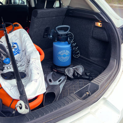 Photography of a white car trunk packed with beach and diving gear, featuring a blue Beach Soul portable shower, a black snorkel mask and grey fins, and an orange diving buoy with a black spearfishing gun
